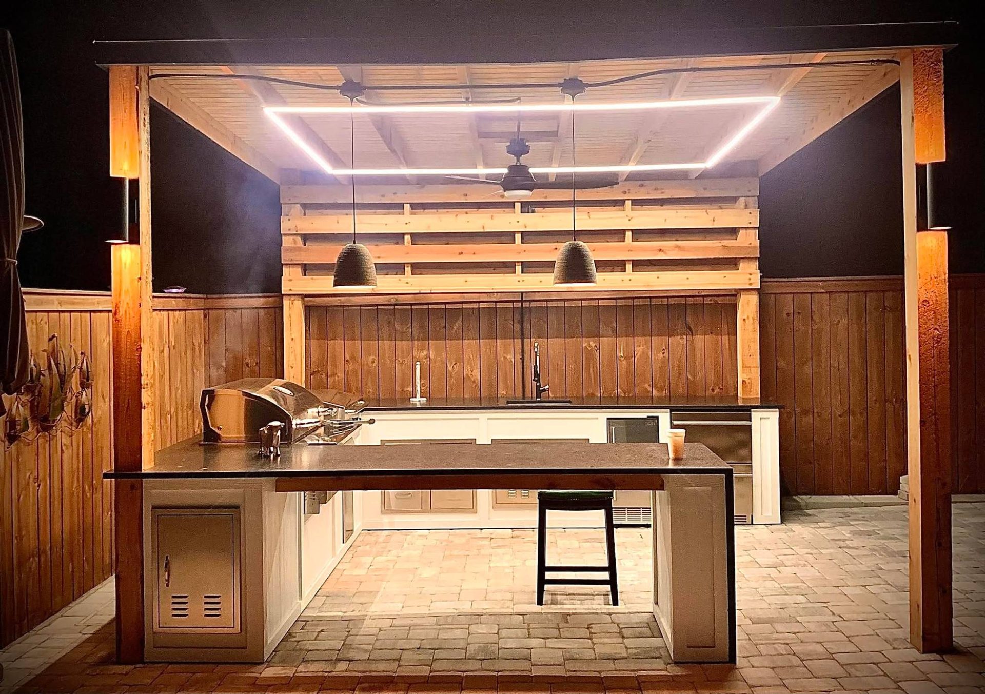 Glowing LED-lined ceiling frame and pendant lights over an outdoor kitchen bar at night
