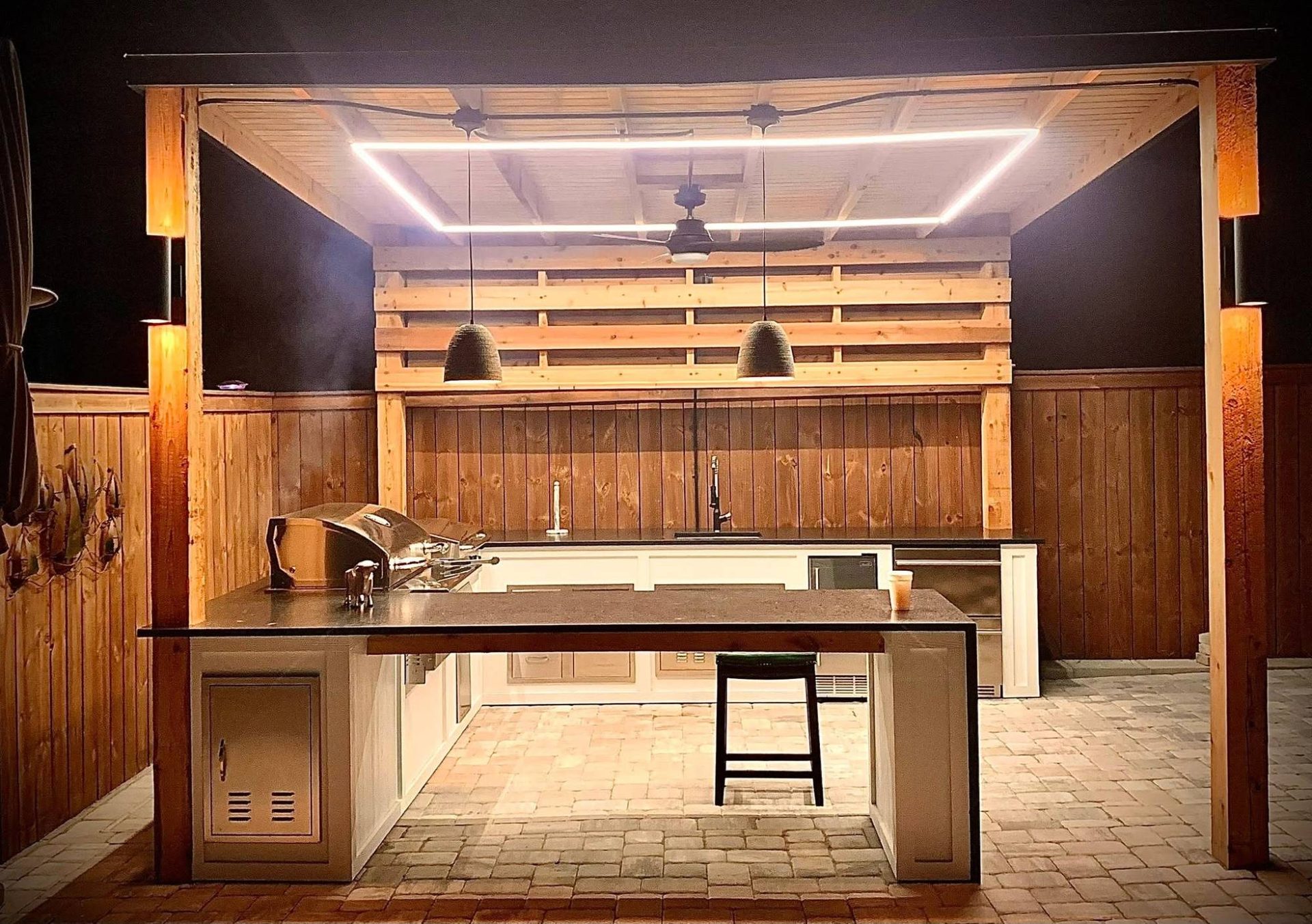 Custom outdoor kitchen under a cedar pergola wired with a glowing LED ceiling frame, pendant lights, and a fan